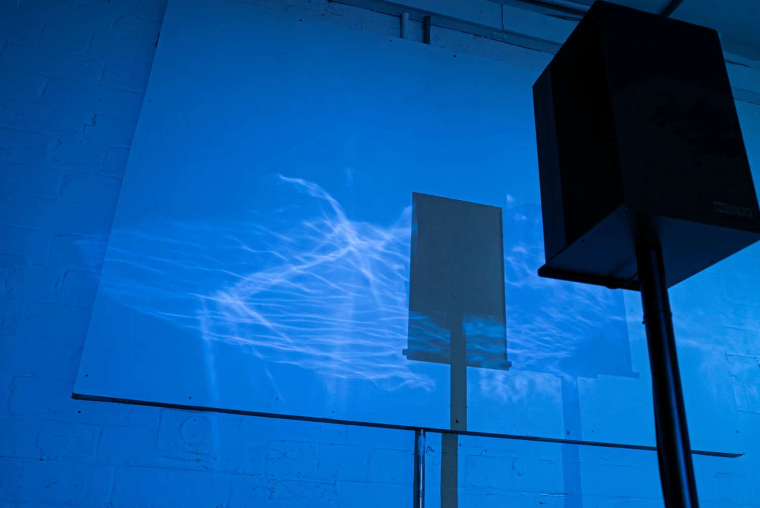 Installation view, water caustics reflection on a white wall behind a large speaker, NCAD Works 2026.